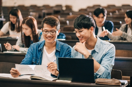 Two male students sitting at a desk in a lecture theatre, smiling and looking towards a piece of paper held by one student