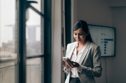 A female student standing in front of a window in an office building, looking downwards towards a phone in her hand