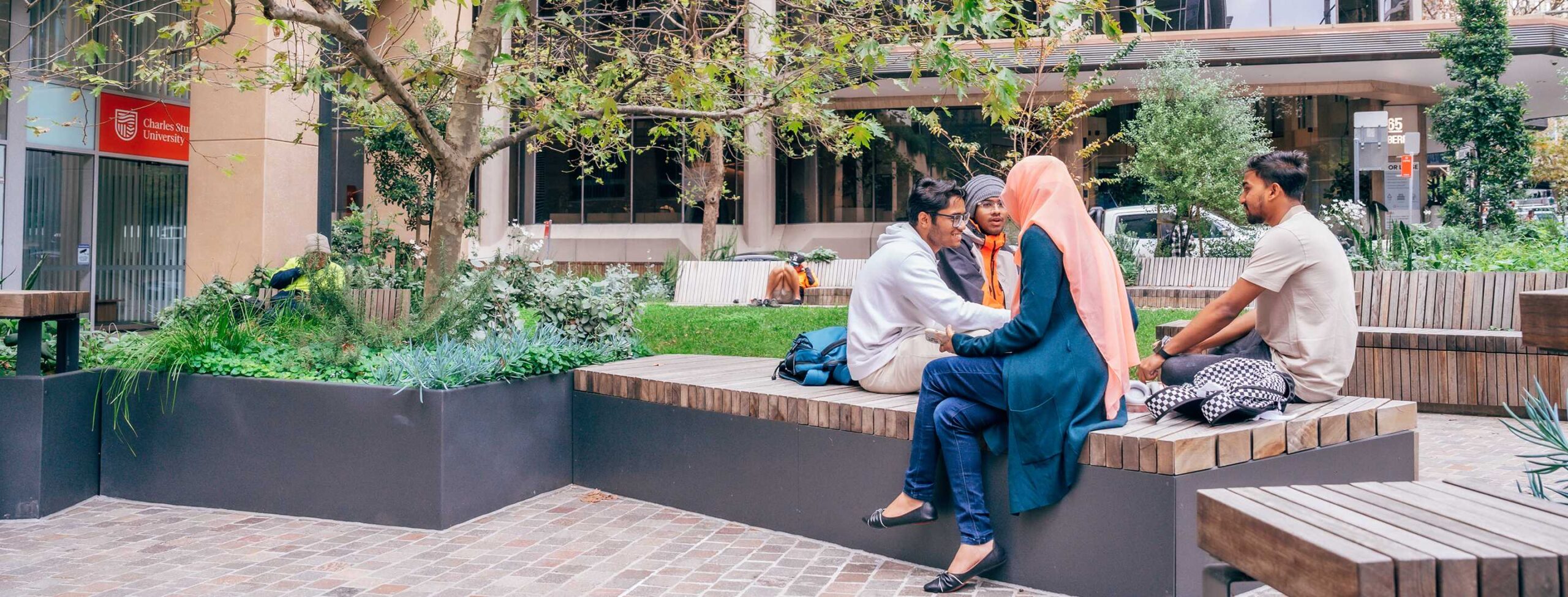 A group of four students sitting and talking on a bench in a garden area outside of Charles Sturt University North Sydney campus