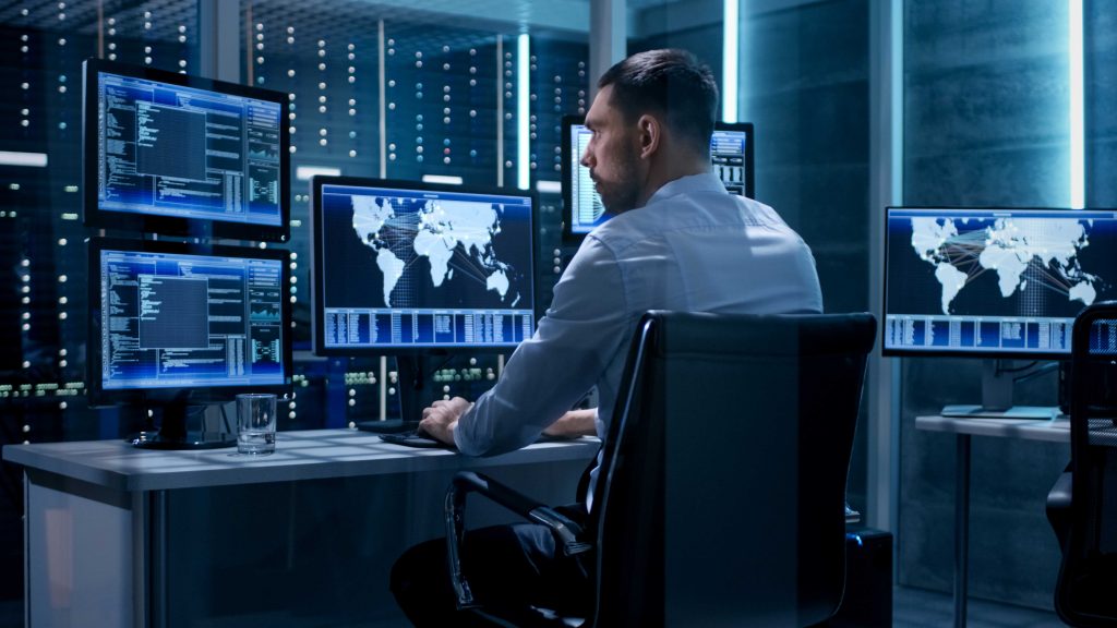 A man sitting in a blue lit room in front of several desktop monitors displaying software engineering tools